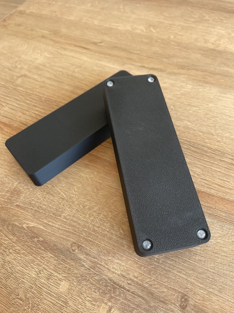 May include: Two black rectangular boxes with rounded edges and silver screws on the bottom.