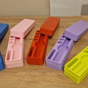 Op de afbeelding: Een verzameling kleurrijke rechthoekige etuis in blauw, roze, rood, paars en geel. Elke etui heeft een bijpassende deksel en een interieurcompartiment met verschillende sleuven en vormen voor het bewaren van pennen en andere benodigdheden.