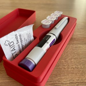 May include: A red plastic case with a compartment for a pen-like medical device, a small packet of alcohol swabs, and four small vials. The pen-like device has a purple cap and the text "Once weekly 0783569 3D Viva 1" printed on the side.