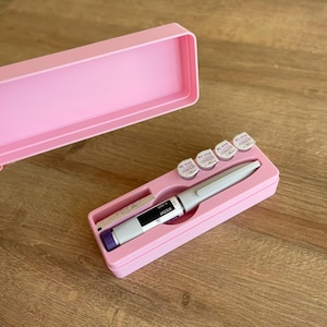 May include: A pink case with a white pen and small round containers. The pen has a purple cap and a label that reads "NovoLog 100". The containers have labels that read "BD Vial 100".