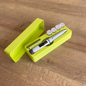 May include: A lime green plastic case with a lid, containing a pen-like medical device and five small white vials with purple labels. The vials have the text "BD Viva 32G 8mm" printed on them.