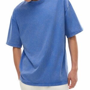 K&ouml;nnte beinhalten: Ein blaues T-Shirt mit kurzen &Auml;rmeln und Rundhalsausschnitt. Das Shirt hat ein verwaschenes, strukturiertes Aussehen. Das Model tr&auml;gt eine wei&szlig;e Hose. Das Shirt ist ein l&auml;ssiges Kleidungsst&uuml;ck f&uuml;r den Alltag.