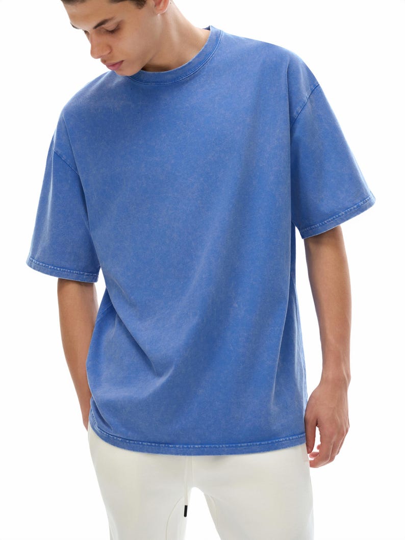 K&ouml;nnte beinhalten: Ein blaues T-Shirt mit kurzen &Auml;rmeln und Rundhalsausschnitt. Das Shirt hat ein verwaschenes, strukturiertes Aussehen. Das Model tr&auml;gt eine wei&szlig;e Hose. Das Shirt ist ein l&auml;ssiges Kleidungsst&uuml;ck f&uuml;r den Alltag.