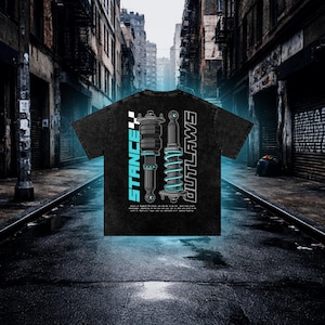 K&ouml;nnte beinhalten: Schwarzes T-Shirt mit einem grafischen Design von Automobil-Federungskomponenten. Die W&ouml;rter "STANCE" und "OUTLAWS" sind vertikal mit einer karierten Flagge dargestellt. Das Design ist in Blau und Wei&szlig; gehalten. Das T-Shirt hat einen Vintage-Look.