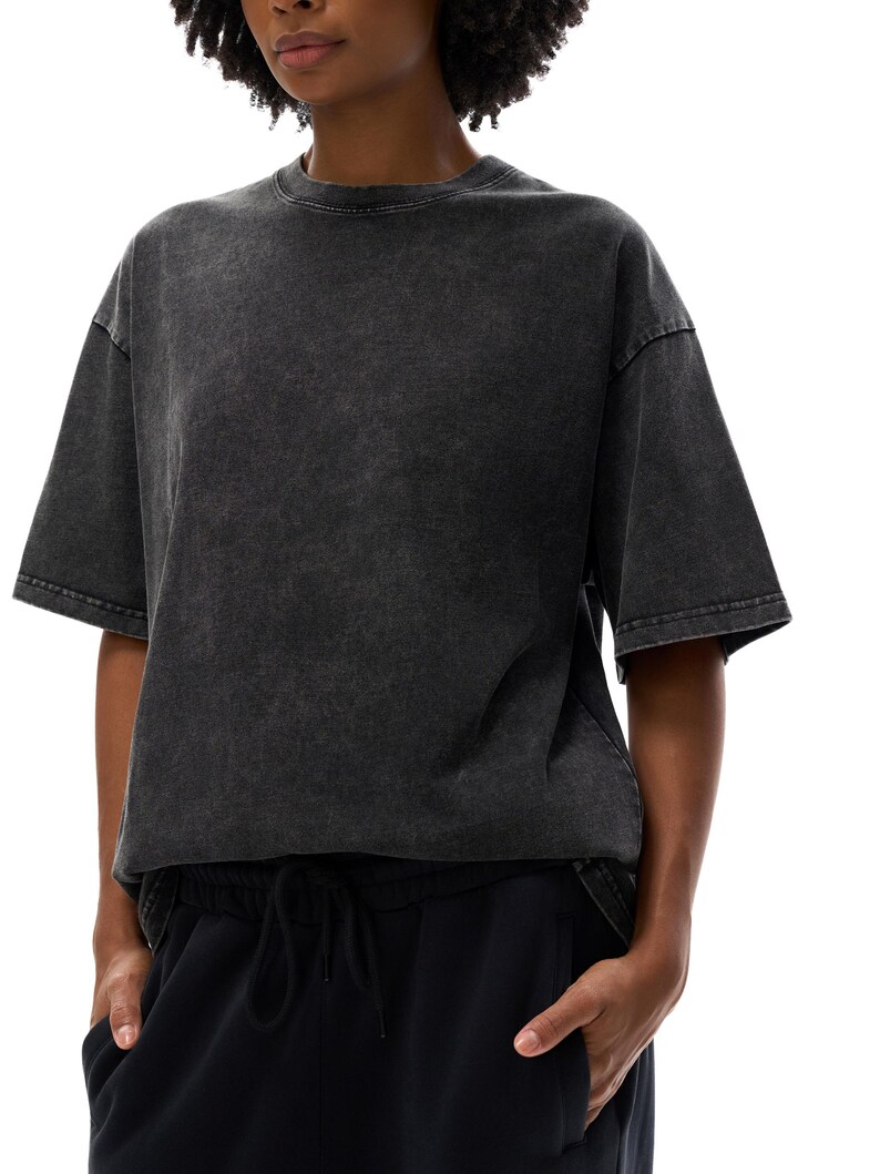 K&ouml;nnte beinhalten: Ein dunkelgraues T-Shirt mit S&auml;urewaschung und kurzen &Auml;rmeln. Das Shirt hat einen Rundhalsausschnitt und eine leicht &uuml;bergro&szlig;e Passform. Das Model tr&auml;gt eine schwarze Hose mit Kordelzug. Der Hintergrund ist wei&szlig;.