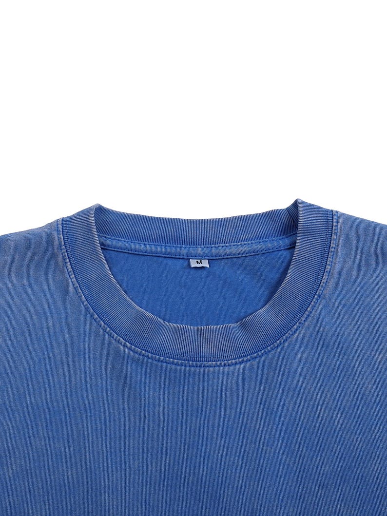 K&ouml;nnte beinhalten: Nahaufnahme eines blauen Rundhals-T-Shirts. Das Shirt hat einen gerippten Kragen und einen verblichenen, Vintage-Look. Das Etikett lautet "M". Der Stoff scheint eine weiche Baumwollmischung zu sein. Das Shirt ist einfarbig und hat einen l&auml;ssigen Stil.