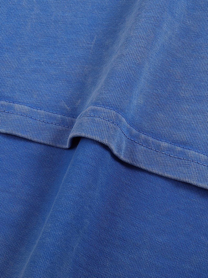 K&ouml;nnte beinhalten: Nahaufnahme eines leuchtend blauen Stoffes, m&ouml;glicherweise ein T-Shirt oder ein anderes Kleidungsst&uuml;ck. Die Textur des Materials ist sichtbar, mit subtilen Farbvariationen und einem markanten Nahtdetail. Der Stoff wirkt weich und leicht abgenutzt.