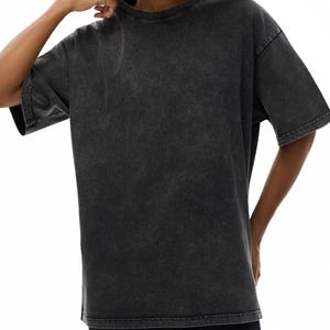 K&ouml;nnte beinhalten: Ein &uuml;bergro&szlig;es T-Shirt in Anthrazit mit Rundhalsausschnitt und kurzen &Auml;rmeln. Das Shirt hat ein verwaschenes, Vintage-Aussehen. Das Model tr&auml;gt das Shirt, die Hand in der N&auml;he des Kragens.