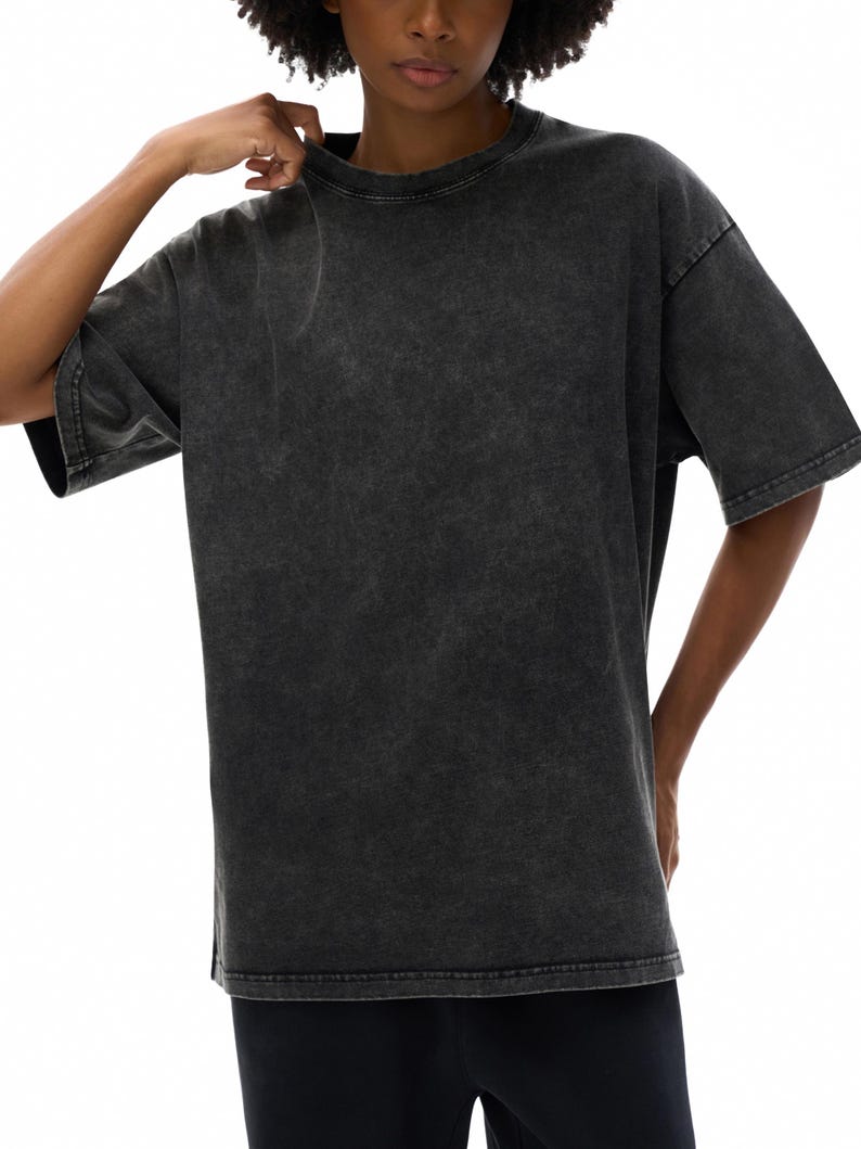 K&ouml;nnte beinhalten: Ein &uuml;bergro&szlig;es T-Shirt in Anthrazit mit Rundhalsausschnitt und kurzen &Auml;rmeln. Das Shirt hat ein verwaschenes, Vintage-Aussehen. Das Model tr&auml;gt das Shirt, die Hand in der N&auml;he des Kragens.