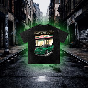 May include: A black acid-wash t-shirt featuring a graphic print of a green sports car parked in front of a JDM STORE. The text MIDNIGHT MEET and Japanese characters are printed above the store illustration, which includes a VIP PARKING ONLY sign.