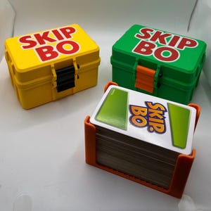 Peut inclure: Trois jeux de cartes Skip-Bo. Les ensembles comprennent des boîtes en plastique jaunes et vertes avec le nom du jeu en lettres rouges. Un jeu de cartes est maintenu dans un support orange. Les cartes ont des motifs verts et blancs.