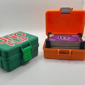 Pode incluir: Duas caixas de plástico para o jogo de cartas UNO FLIP. Uma caixa verde está fechada, com o título do jogo em vermelho. A outra é laranja e está aberta, revelando um baralho de cartas. As caixas têm suportes de cartas pretos.