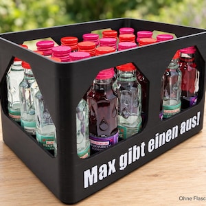 Op de afbeelding: Een zwarte kunststof drankkrat met meerdere vakken met glazen flessen met felrode doppen. De zijkant van de krat is voorzien van de witte tekst "Max gibt einen aus!" en in de onderste hoek staat de tekst "Ohne Flaschen & Inhalt".