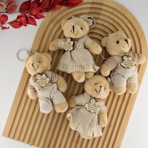 Puede incluir: Cinco llaveros de osos de peluche. Los osos son de color beige con ropa de tela en tonos neutros. Cada oso tiene una etiqueta de madera con texto. Los llaveros están colocados en una bandeja de madera arqueada, con follaje rojo en el fondo.