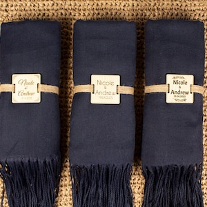 Könnte beinhalten: Drei marineblaue Pashmina-Schals mit Holzetiketten, auf denen "Nicole & Andrew" mit dem Hochzeitsdatum steht. Die Schals liegen auf einem gewebten Strohhintergrund.