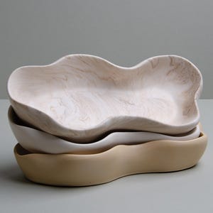 May include: Three ceramic bowls in different sizes and colors, stacked on top of each other. The top bowl is white with a light brown marbled pattern, the middle bowl is white, and the bottom bowl is a light beige color.