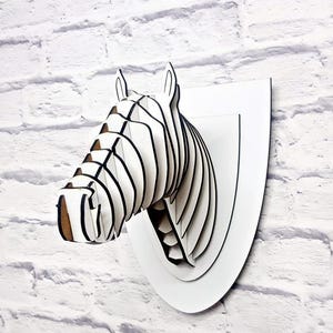 Puede incluir: Una cabeza de cebra de madera blanca montada en una placa blanca. La cabeza de cebra está hecha de madera en capas y tiene un patrón de rayas blancas y negras.