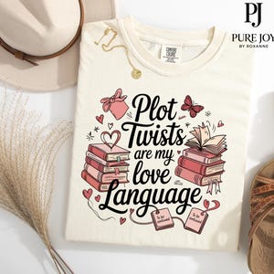May include: A white t-shirt with a pink and black graphic design featuring a stack of books, a butterfly, hearts, and the text "Plot Twists are my love Language".