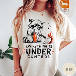 May include: An ivory t-shirt featuring a graphic of a raccoon holding an orange fire extinguisher and spraying a burning to-do list. The text below the illustration reads, "EVERYTHING IS UNDER CONTROL" in bold black letters with orange accents.