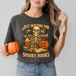 Puede incluir: Camiseta gris oscuro con un diseño temático de Halloween. El diseño presenta un esqueleto, una calabaza, libros y el texto "Just Waiting For Spooky Books". Una taza naranja con forma de calabaza se sostiene en la mano.