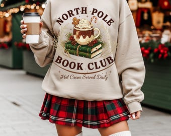 Könnte beinhalten: Beiges Sweatshirt mit einem Aufdruck "North Pole Book Club" mit einer heißen Schokoladentasse, einem Lebkuchenmann und einem Zuckerstangen-Design. Darunter steht "Hot Cocoa Served Daily". Die Person trägt einen rot-grünen karierten Rock.