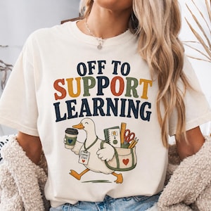 Puede incluir: Una camiseta de color crema con el texto OFF TO SUPPORT LEARNING en azul oscuro, naranja y verde. Debajo del texto hay un gráfico de un pato blanco que sostiene una taza de café y una bolsa con un cuaderno, tijeras y lápices.