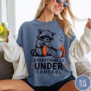 May include: A blue t-shirt featuring a graphic of a raccoon holding an orange fire extinguisher and spraying a burning to-do list. The text below the graphic reads, "EVERYTHING IS UNDER CONTROL" in black capital letters with orange accents.