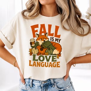 May include: A cream-colored t-shirt featuring a graphic of a fox wearing a green sweater, holding a coffee cup, and surrounded by pumpkins and autumn leaves. The text reads "FALL IS MY LOVE LANGUAGE" in orange and green block lettering.