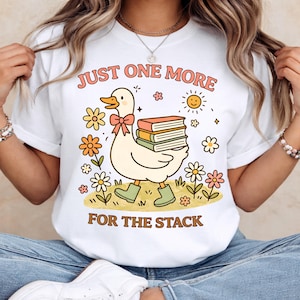Puede incluir: Camiseta blanca con un diseño caprichoso de un pato con botas verdes y un lazo, llevando libros. El texto "JUST ONE MORE FOR THE STACK" está en una fuente retro, rodeado de flores y un sol sonriente.
