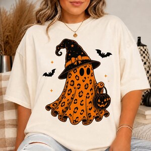 May include: An off-white t-shirt featuring a graphic of a ghost wearing a black witch hat with an orange bow. The ghost has an orange leopard print pattern and is holding a small orange jack-o'-lantern bucket. Two black bats and small orange stars surround the ghost.