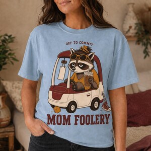 May include: A light blue t-shirt featuring a graphic of a raccoon wearing sunglasses and a crown while driving a golf cart. The raccoon holds a drink, and the text reads OFF TO COMMIT MOM FOOLERY in dark red lettering.