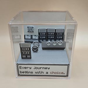 May include: A clear acrylic cube featuring a pixelated diorama scene. The scene includes a character and a building interior with shelves and a counter. The text "Every journey begins with a choice" is displayed on a white label at the bottom.