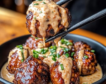 May include: A bowl of savory meatballs in a creamy sauce, topped with fresh green onions. The meatballs are dark brown and have a slightly crispy exterior. The sauce is a light brown color and has a smooth, creamy texture.