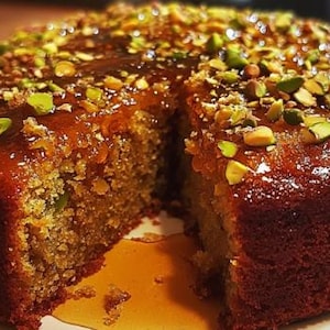 May include: A close-up of a round cake with a slice removed, revealing a moist interior. The cake is topped with a glossy glaze and sprinkled with chopped pistachios. The cake sits on a white plate, with some glaze dripping down the side.