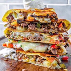 Puede incluir: Una pila de cuatro quesadillas rellenas de queso derretido, carne, pimientos y cebollas. Las quesadillas son doradas y crujientes, y están cubiertas con una cucharada de crema agria.