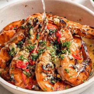May include: A white bowl filled with grilled prawns, close-up shot. The prawns are a vibrant orange, drizzled with sauce and garnished with chopped green herbs and red peppers. The bowl is set against a neutral background.