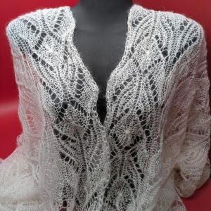 May include: A white lace shawl with a delicate, intricate pattern. The shawl is draped over a mannequin and has a V-neckline.