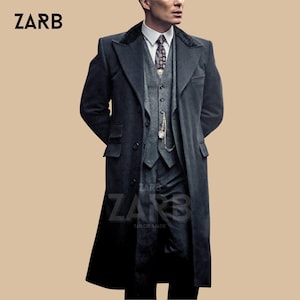 Puede incluir: Un traje de tres piezas gris oscuro a medida con un abrigo largo. El traje incluye un chaleco, un pantalón y una camisa con cuello y corbata estampada. Un reloj de bolsillo cuelga del chaleco. La marca "ZARB" es visible.