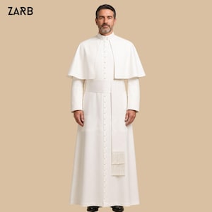 May include: A white cassock with a matching shoulder cape and a wide sash featuring a fringed end. The garment has a high collar, long sleeves, and a vertical row of small buttons running down the center front. The text ZARB is visible in the corner.