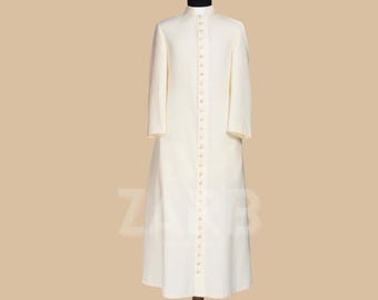 Può includere: Una veste bianca a maniche lunghe con chiusura a bottoni e colletto alto. La veste è su un manichino di legno.