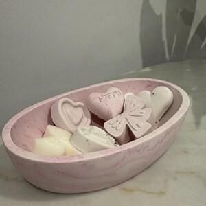 May include: A pink oval bowl containing heart, butterfly, and round wax melts in shades of pink and white. One heart-shaped melt has the word "HAPPY" on it. The bowl has a marbled effect. The bowl is on a marble surface.