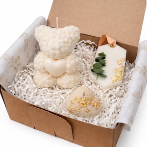 May include: A gift box with three ivory candles. One is a teddy bear shape, another a tag with greenery, and the third a heart shape with gold leaf. The box is lined with paper and decorative tissue.