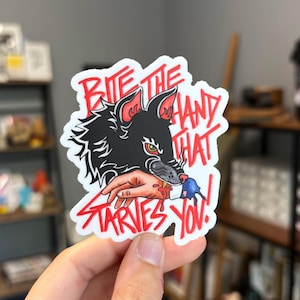 May include: A sticker featuring a black wolf biting a hand with red blood dripping. The text "Bite the hand that starves you!" is written in red, stylized lettering. The sticker has a white border.