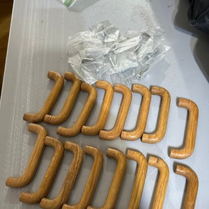 Puede incluir: Un conjunto de tiradores de armario de madera con un diseño curvo en forma de D. Los tiradores son de color marrón claro y parecen ser de madera. También se ven pequeñas bolsas de ferretería.