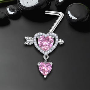 Könnte beinhalten: Ein silberfarbener Bauchnabelpiercing mit einem herzförmigen rosa Edelstein, der von kleinen, klaren Steinen umgeben ist. Ein Pfeildesign durchbohrt das Herz, und ein kleineres Herz hängt darunter.