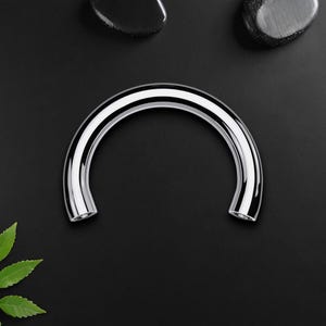 Könnte beinhalten: Ein glänzendes, gebogenes Metallobjekt mit polierter Chromoberfläche. Das Objekt hat eine abgerundete, halbkreisförmige Form mit hohlem Innenraum. Der Hintergrund ist schwarz, mit ein paar dunklen Steinen und grünen Blättern.