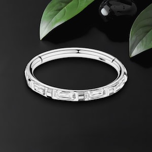 May include: A silver bangle bracelet with rectangular, clear gemstones set around the band. The bracelet is displayed on a black surface, with green leaves and a black stone in the background.