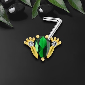 Puede incluir: Un piercing de nariz con una gran piedra preciosa verde esmeralda engastada en un marco dorado. El marco tiene un diseño estilizado con pequeñas piedras de acento transparentes. El poste es plateado y está doblado en ángulo recto.