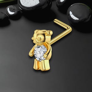 May include: A gold-colored nose ring in the shape of a teddy bear. The bear has a clear, round gemstone in its belly. The nose ring is on a black background with black, glossy stones.