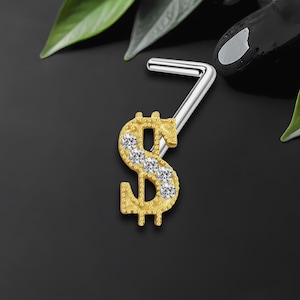 May include: A gold-colored nose stud in the shape of a dollar sign, embellished with clear, sparkling gemstones. The stud has a silver-colored, L-shaped post. The background is dark, with a few green leaves and a black, rounded object.